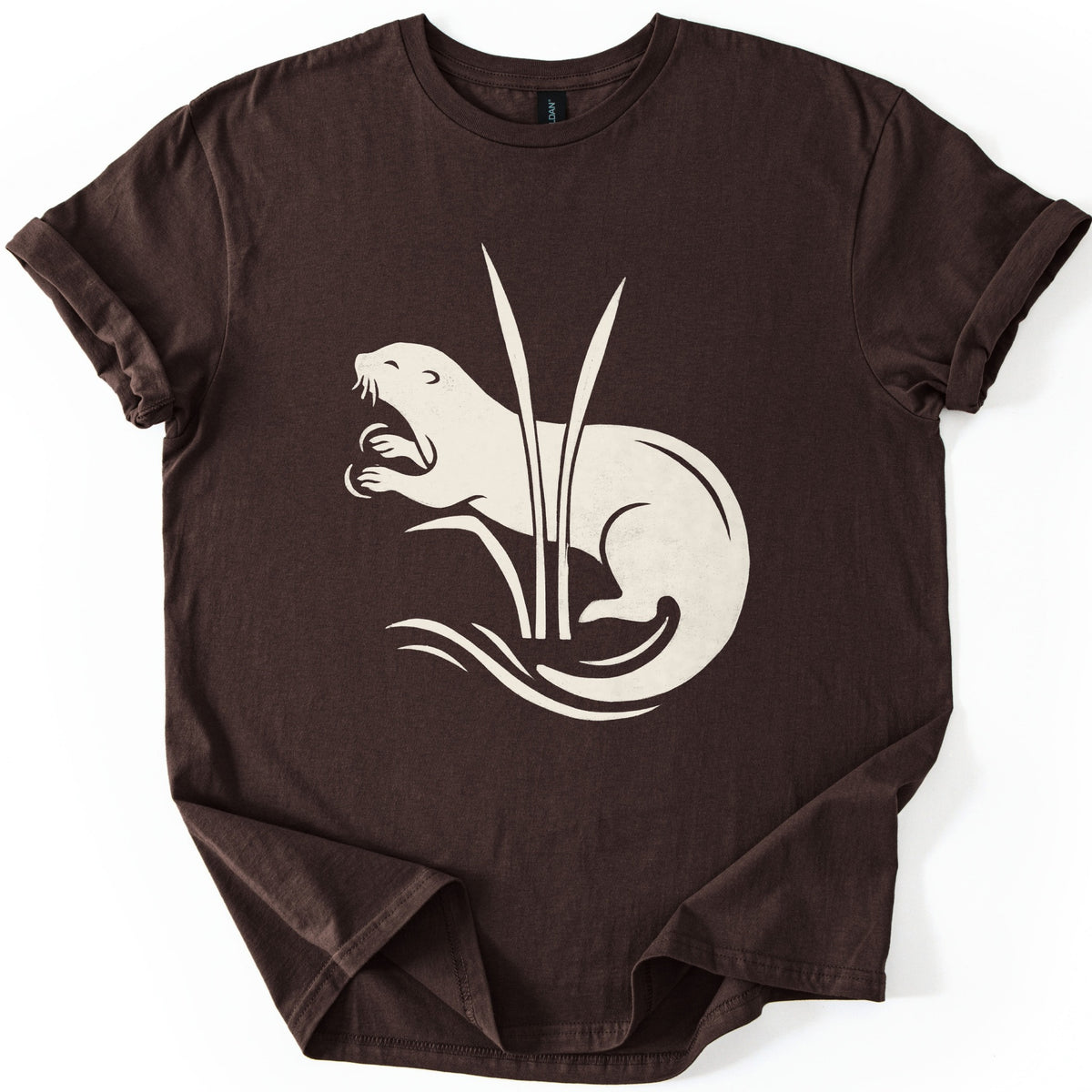 Otter In The Reeds Comfort Colors T-shirt in Dark Chocolate, laid flat. A cream silhouette of an otter with open mouth, whiskers and raised paws curls behind two tall reeds above swirling water lines. No words. The shirt lies on a white surface with sleeves rolled.