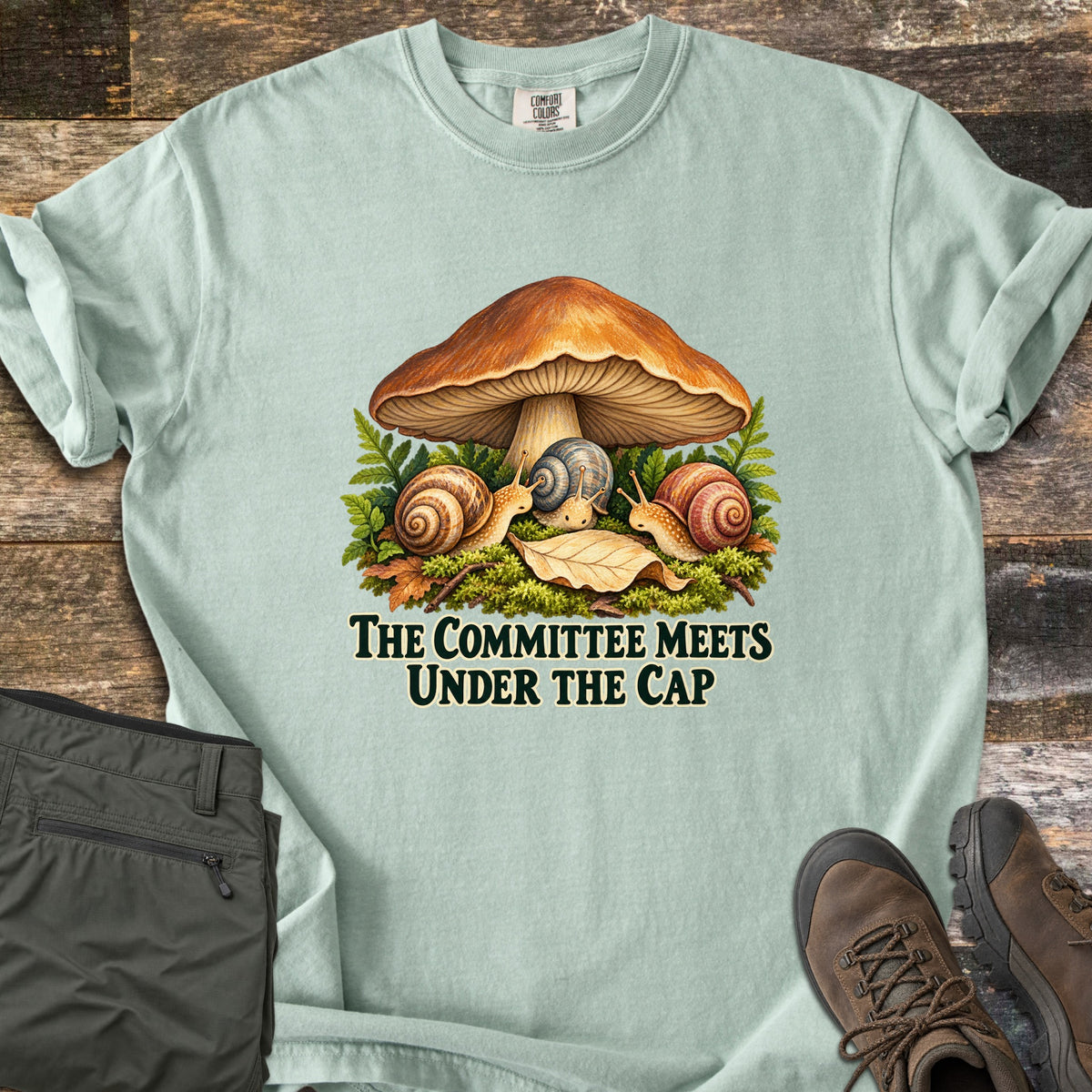 The Committee Meets Under the Cap Comfort Colors T-shirt in Bay, laid flat. A large brown mushroom shelters three snails on moss, ferns and fallen leaves above the words The Committee Meets Under the Cap in dark green. Olive pants and brown hiking boots sit on a weathered wood surface.