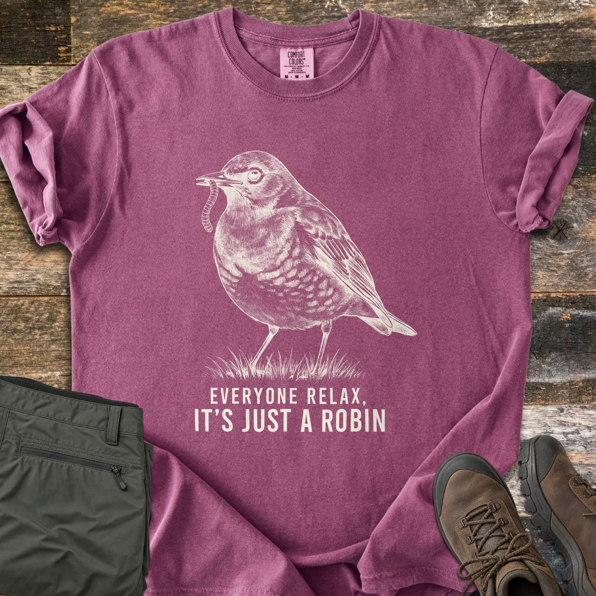 Everyone Relax It's Just A Robin Comfort Colors T-shirt in Berry, laid flat. A sketched robin with a worm in its beak stands on grass above the words Everyone Relax, It's Just A Robin, in cream ink. Olive cargo pants and brown hiking boots lie beside it on weathered wood.