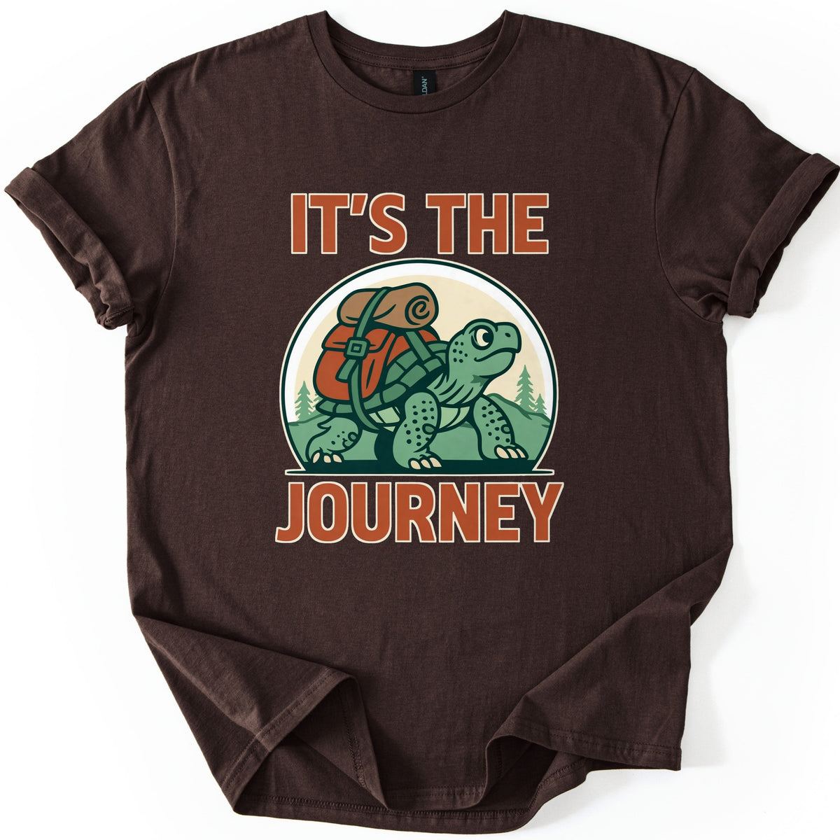 Slow Turtle Hiker Journey Comfort Colors T-shirt in Dark Chocolate, laid flat. A green cartoon turtle carrying a red backpack with a rolled sleeping bag walks past pine trees, framed by the words It's the Journey in rust-orange letters. Sleeves are cuffed on a white background.
