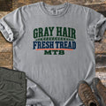 Gray Hair Fresh Tread Comfort Colors T-shirt in Grey, laid flat. Arched green text reads GRAY HAIR over a green tire tread band, then navy FRESH TREAD, an orange line, and green MTB. Olive hiking pants and brown hiking boots sit beside it on weathered wood planks.