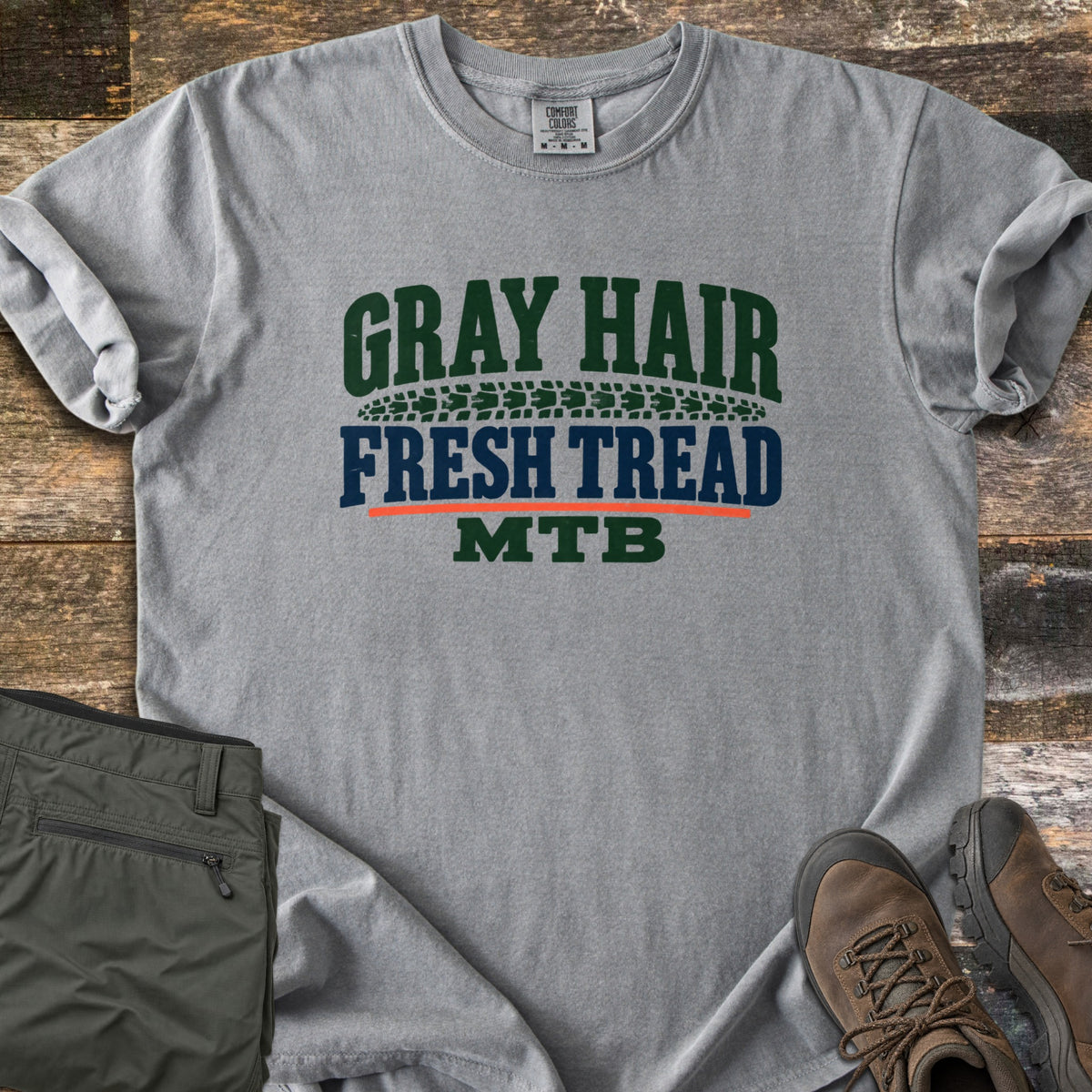 Gray Hair Fresh Tread Comfort Colors T-shirt in Grey, laid flat. Arched green text reads GRAY HAIR over a green tire tread band, then navy FRESH TREAD, an orange line, and green MTB. Olive hiking pants and brown hiking boots sit beside it on weathered wood planks.