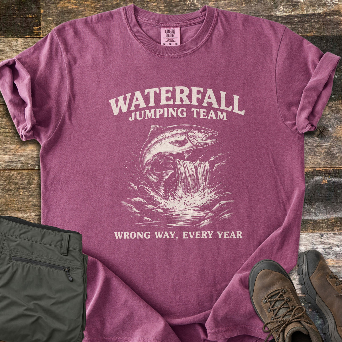 Waterfall Jumping Team Comfort Colors T-shirt in Berry, laid flat. A salmon leaps beside a small waterfall between the words Waterfall Jumping Team and Wrong Way, Every Year, printed in cream. Shown on rustic wood with folded olive pants and brown hiking boots.