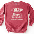 Opossum Acting School Comfort Colors crewneck sweatshirt in Crimson, laid flat. Cream print of a sketched opossum lying on its back in leaves, mouth open, under the words Opossum Acting School and above Major: Playing Dead. The sweatshirt lies on a white surface.
