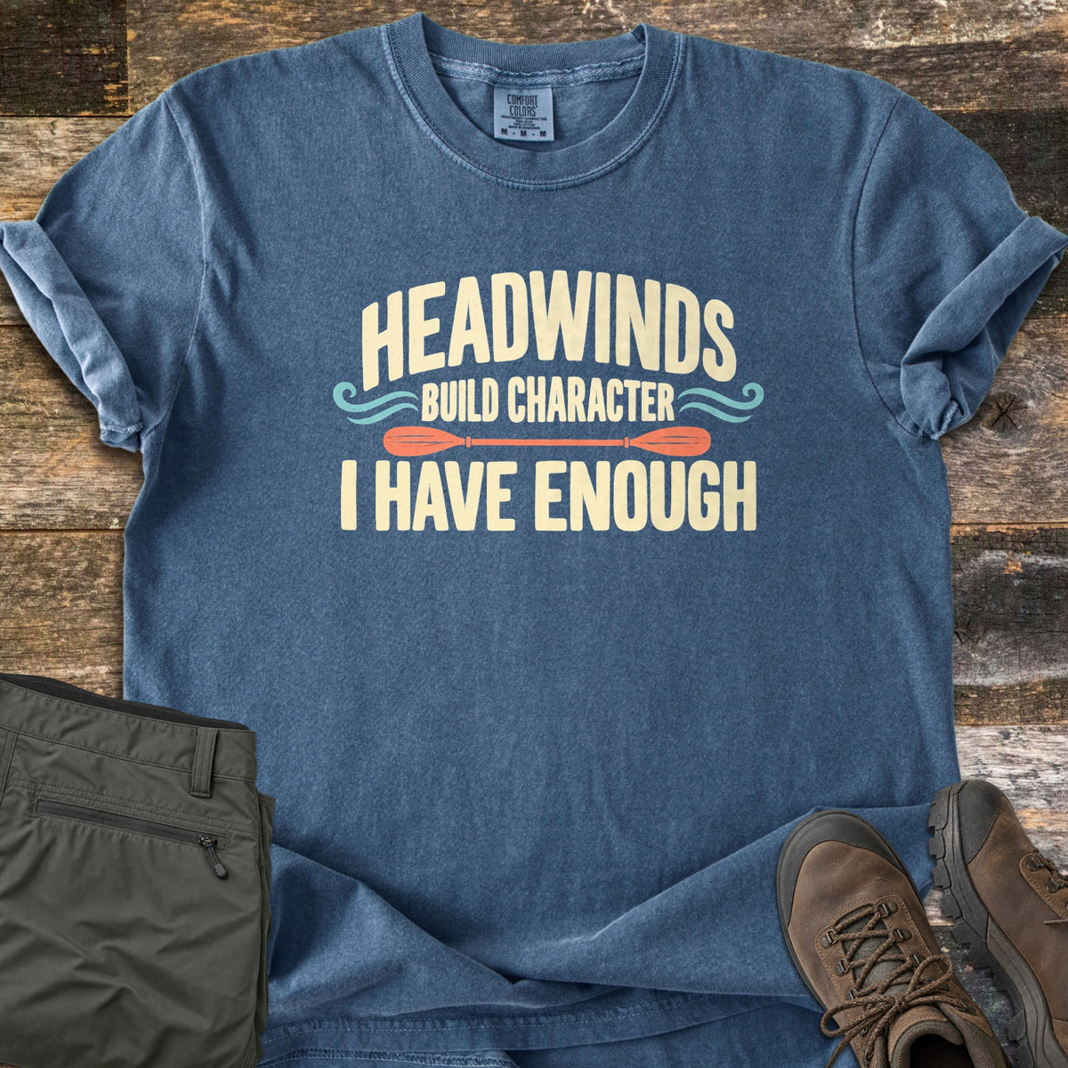 Headwinds Build Character I Have Enough Comfort Colors T-shirt in Denim, laid flat. Cream text reads Headwinds Build Character I Have Enough, with teal wave swirls and an orange kayak paddle. Sleeves rolled, on weathered wood beside olive hiking pants and brown hiking boots.