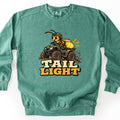 Tail Light Comfort Colors crewneck sweatshirt in Light Green, laid flat. A cartoon firefly in a green helmet rides a muddy green ATV, its yellow tail glowing, above the words Tail Light in cream and yellow block letters. The sweatshirt lies on a white surface.