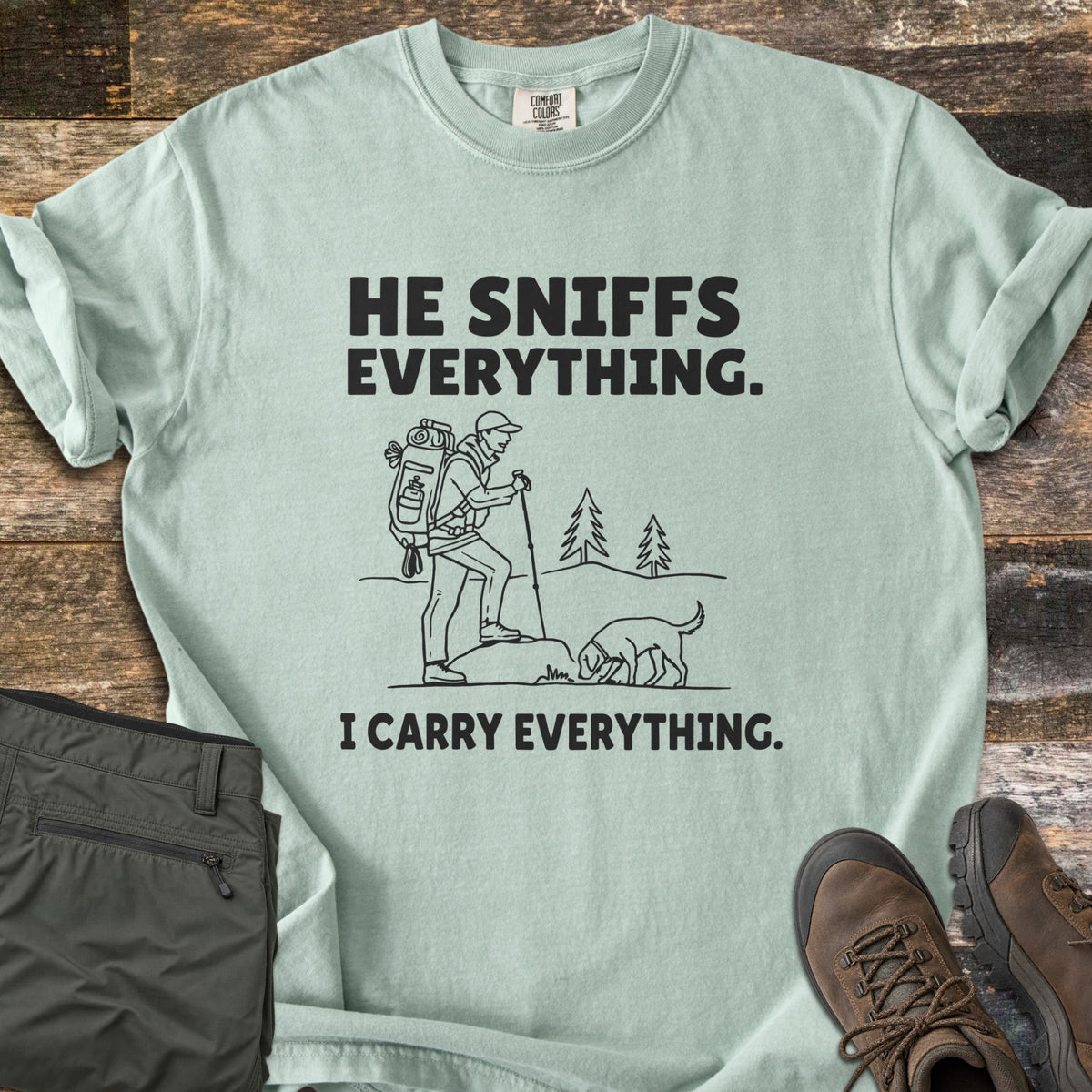 He Sniffs Everything. I Carry Everything Comfort Colors T-shirt in Bay, laid flat. Black line art of a hiker with a large backpack and trekking pole beside a dog sniffing a rock, between the words He Sniffs Everything. and I Carry Everything. Olive pants and brown hiking boots sit on weathered wood.