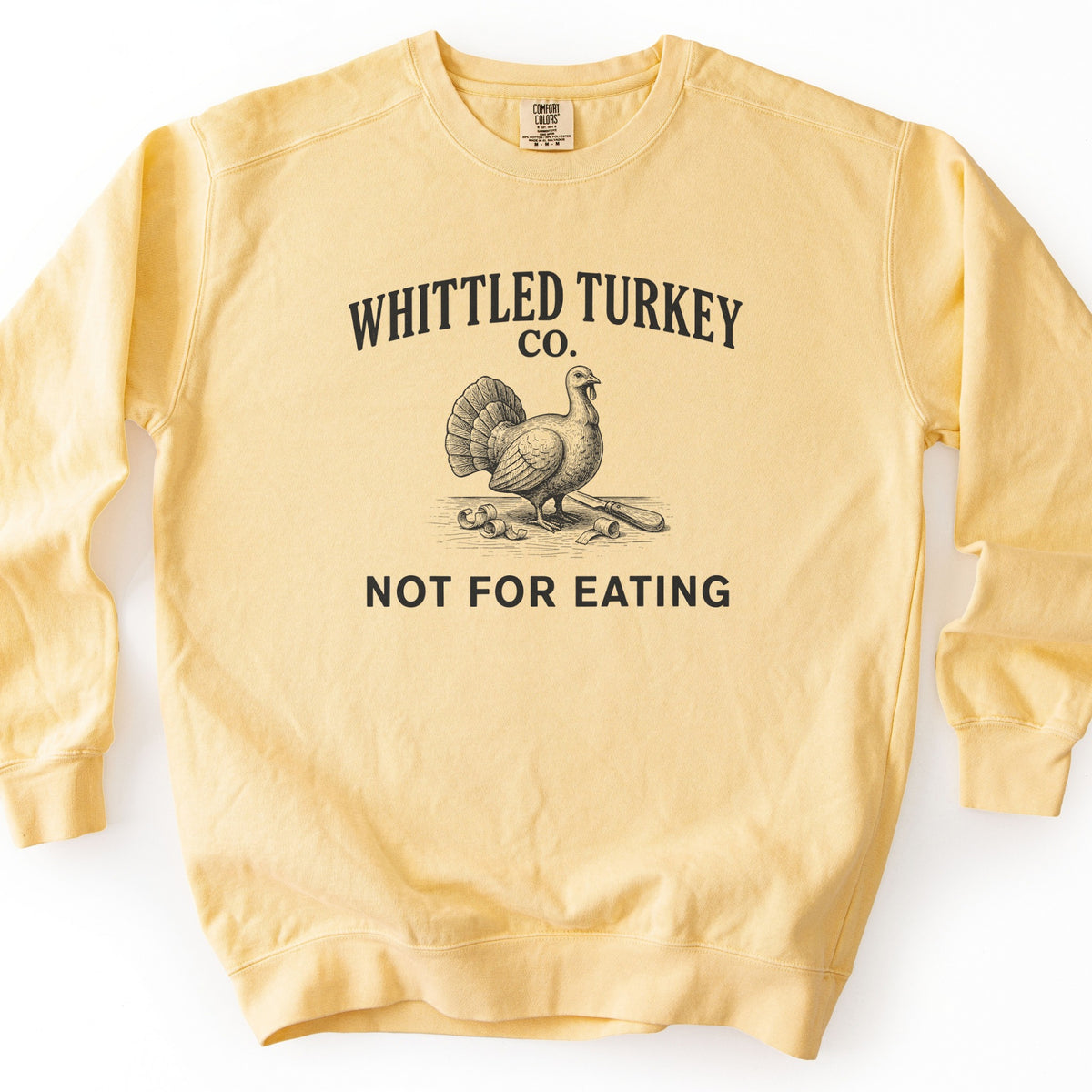 Whittled Turkey Co Comfort Colors crewneck sweatshirt in Butter, laid flat. Black print of the words Whittled Turkey Co. arched over an engraved-style turkey standing beside a carving knife and wood shavings, with Not For Eating below. The sweatshirt lies on a white background.