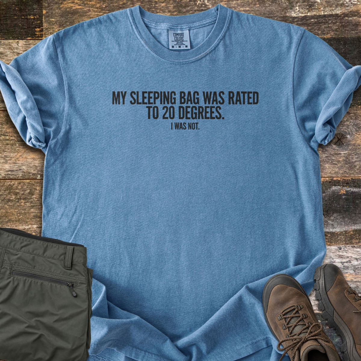 Rated To 20 Degrees Comfort Colors T-shirt in Blue Jean, laid flat. Black text across the chest reads My Sleeping Bag Was Rated To 20 Degrees. I Was Not. The shirt has rolled sleeves and lies on weathered wood planks beside folded olive hiking pants and brown hiking boots.