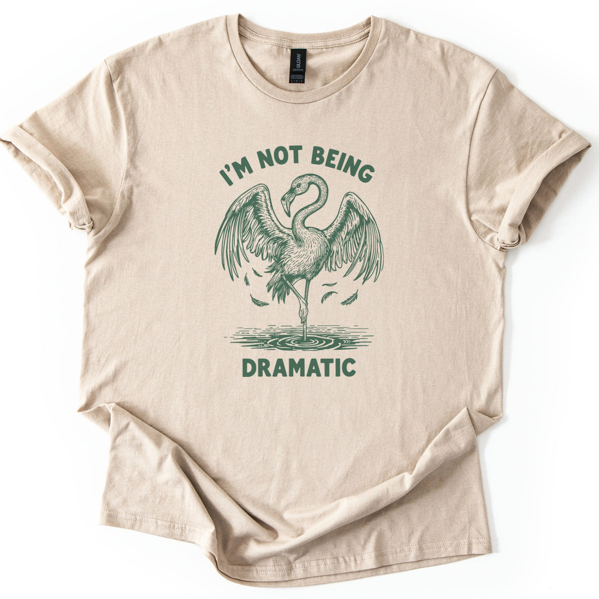I'm Not Being Dramatic Comfort Colors T-shirt in Sand, laid flat. A dark green sketch of a flamingo standing on one leg in rippling water, wings spread wide as feathers fall, sits under the arched words I'm Not Being and above Dramatic. Sleeves are cuffed on a white surface.