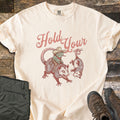 Possum and Frog Comfort Colors T-shirt in Ivory, laid flat. Pink script reads Hold Your above a frog in a cowboy hat and bandana riding a snarling possum toward another possum hanging upside down by its tail. Laid on weathered wood with folded olive pants and brown hiking boots.