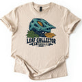 Leaf Collector By Accident Comfort Colors T-shirt in Sand, laid flat. A teal mountain bike helmet with an orange oak leaf in a vent rests on green and gold leaves above the words Leaf Collector By Accident MTB. The shirt lies on a white surface with cuffed sleeves.