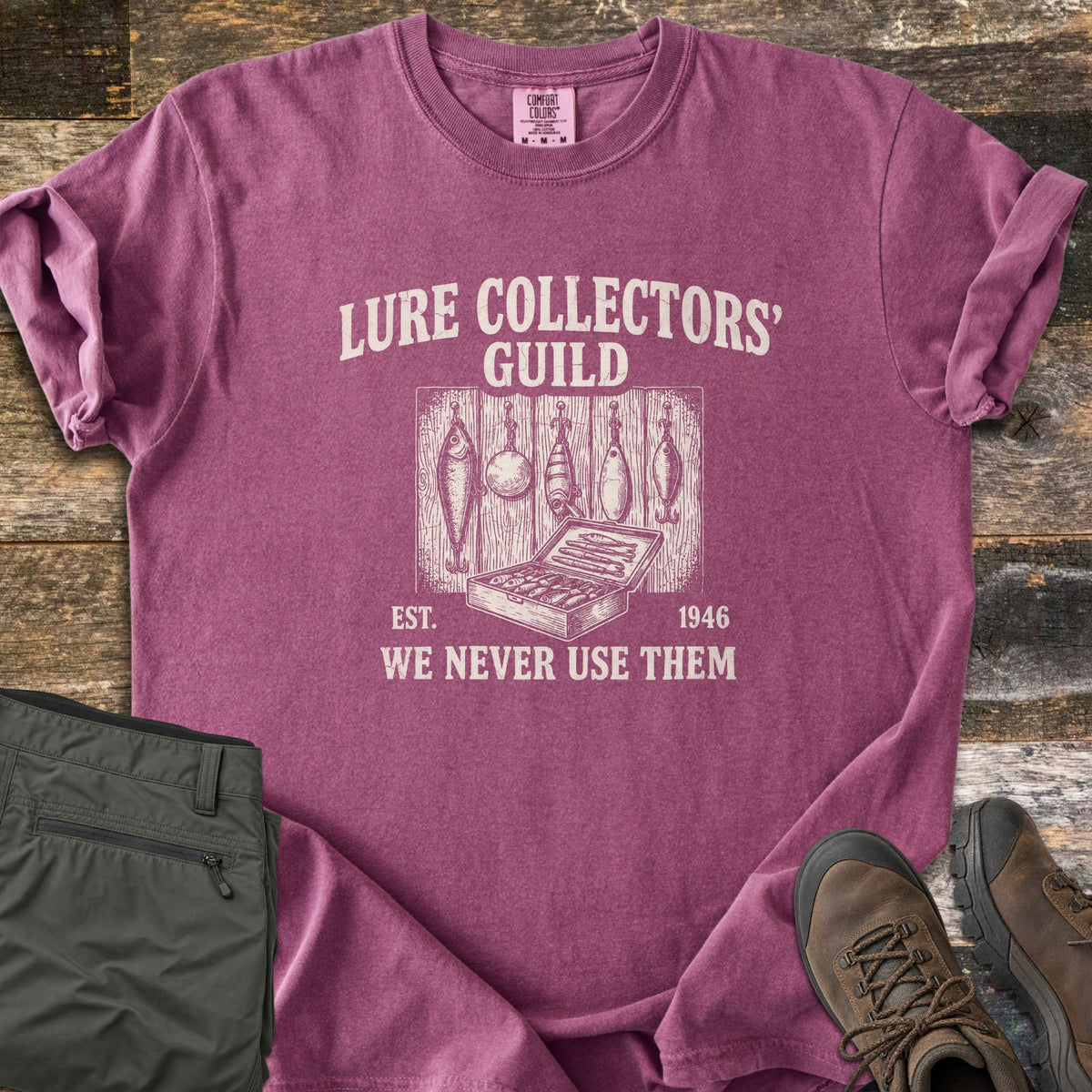 Lure Collectors Guild Comfort Colors T-shirt in Berry, laid flat. A vintage-style drawing of five fishing lures hanging on wood planks above an open tackle box, framed by Lure Collectors' Guild, Est. 1946, We Never Use Them. Olive pants and brown hiking boots sit on wood.