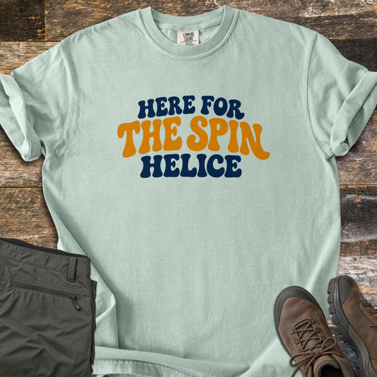 Here for the Spin Comfort Colors T-shirt in Bay, laid flat. Retro wavy lettering reads HERE FOR THE SPIN HELICE, with HERE FOR and HELICE in navy and THE SPIN in gold. The shirt lies on weathered wood with folded olive pants and brown hiking boots.