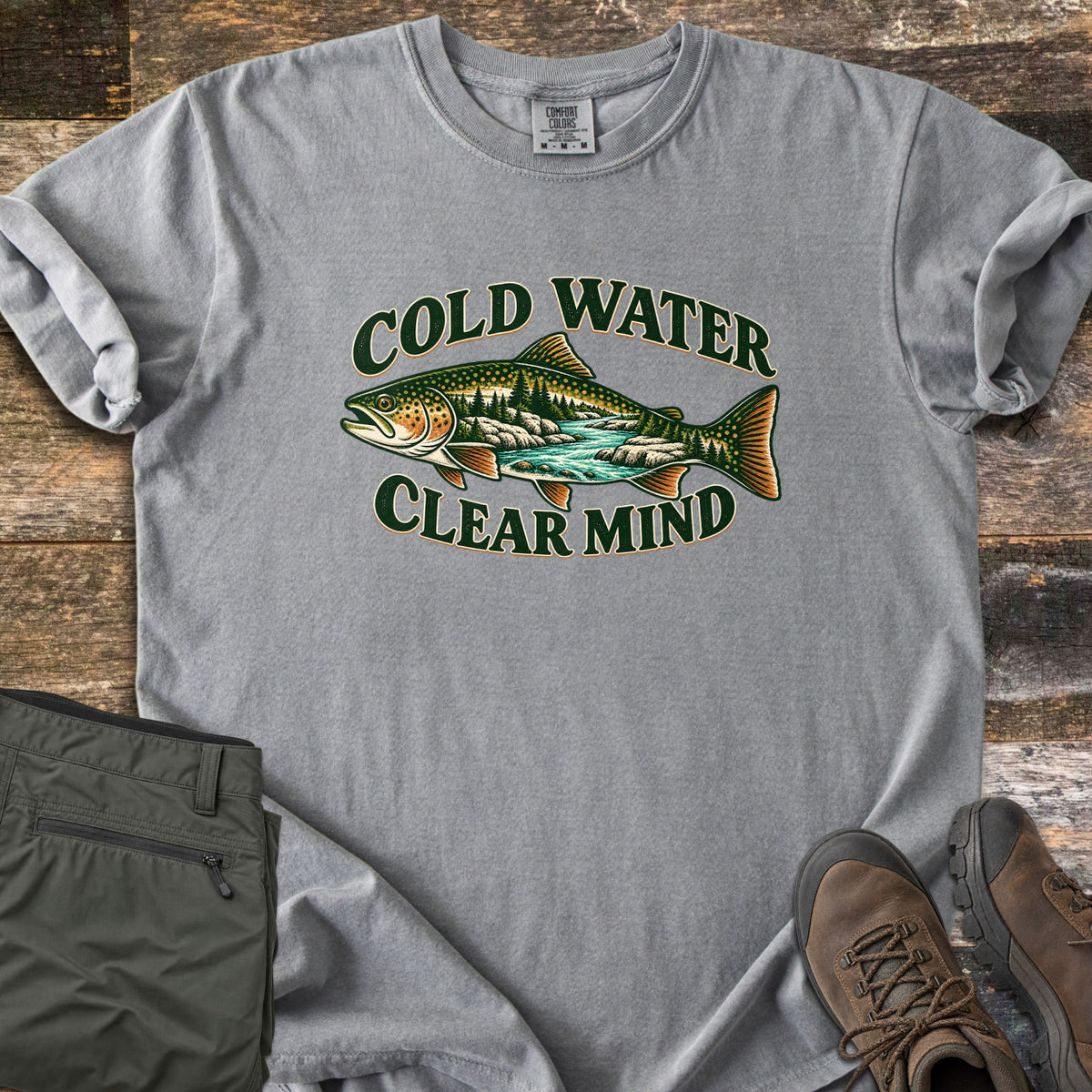 Cold Water Clear Mind Comfort Colors T-shirt in Grey, laid flat. A spotted trout with a pine forest and rushing turquoise river inside its body sits between the dark green words Cold Water and Clear Mind. Folded olive hiking pants and brown hiking boots lie on weathered wood.