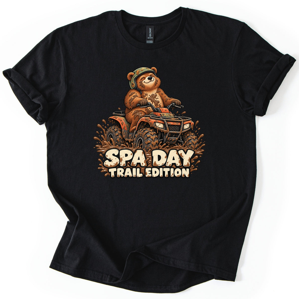 Spa Day Trail Edition Comfort Colors T-shirt in Black, laid flat. A smiling cartoon brown bear in a camo helmet rides a mud-covered orange ATV through splashing mud above the words SPA DAY TRAIL EDITION. The shirt lies on a white surface with rolled sleeves.