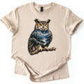 Owl Mountain Lake Comfort Colors T-shirt in Sand, laid flat. A great horned owl with yellow eyes perches on a branch, its body filled with a night scene of a crescent moon, mountains, pine trees and a lake. No words are printed. The shirt lies on a white background.