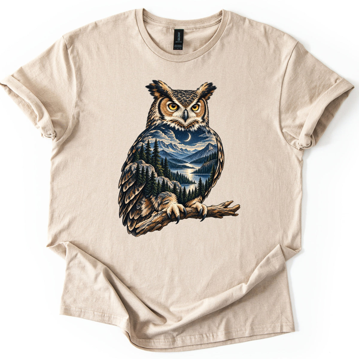 Owl Mountain Lake Comfort Colors T-shirt in Sand, laid flat. A great horned owl with yellow eyes perches on a branch, its body filled with a night scene of a crescent moon, mountains, pine trees and a lake. No words are printed. The shirt lies on a white background.