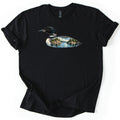 Loon Lake Comfort Colors T-shirt in Black, laid flat. A common loon in profile, its body filled with a lake scene of pine-covered rocky islands, blue water and clouds; no words are printed. The shirt lies on a white surface with its sleeves rolled.