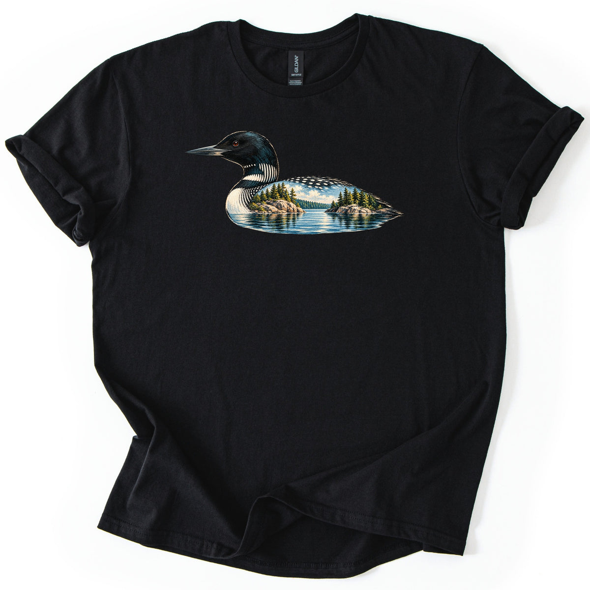 Loon Lake Comfort Colors T-shirt in Black, laid flat. A common loon in profile, its body filled with a lake scene of pine-covered rocky islands, blue water and clouds; no words are printed. The shirt lies on a white surface with its sleeves rolled.