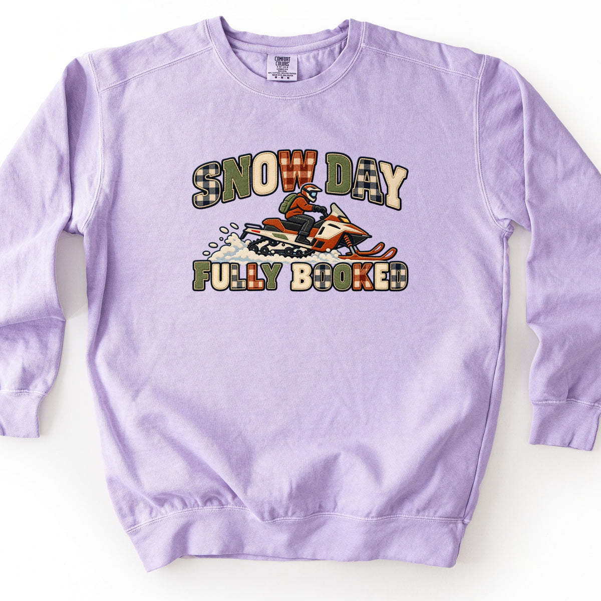 Snow Day Fully Booked Comfort Colors crewneck sweatshirt in Orchid, laid flat. A rider in a helmet and green backpack on an orange and white snowmobile kicks up snow between the words Snow Day and Fully Booked in plaid patch-style letters. Shown on a white surface.