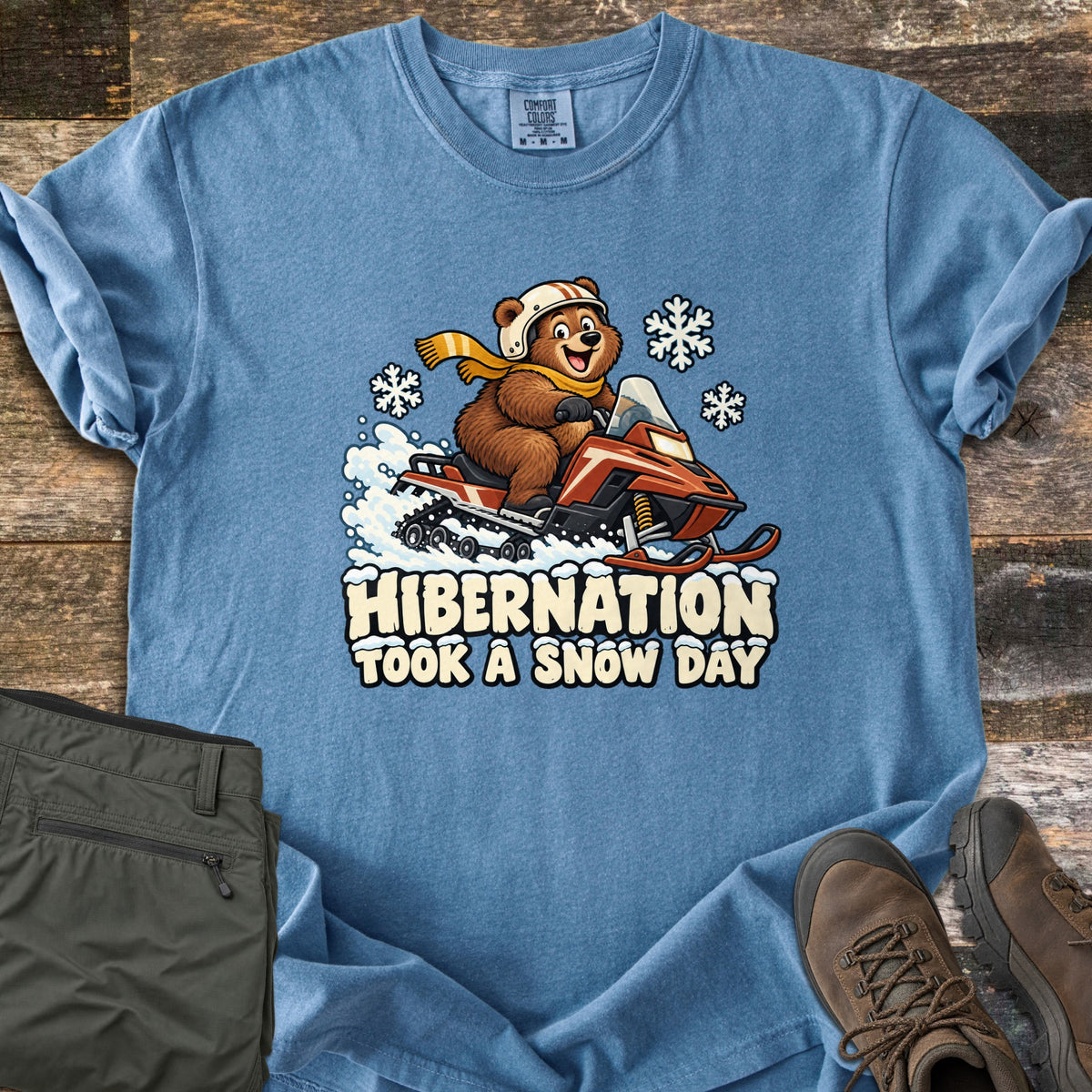 Hibernation Took a Snow Day Comfort Colors T-shirt in Blue Jean, laid flat. A grinning cartoon bear in a white helmet and yellow scarf rides a red snowmobile through snow above the words Hibernation Took a Snow Day. Olive hiking pants and brown boots sit on weathered wood.