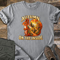 Autumn on the Inside Comfort Colors T-shirt in Grey, laid flat. A red squirrel whose tail frames a forest path through orange and yellow autumn trees, with the words Autumn on the Inside. Olive cargo pants and brown hiking boots lie beside it on weathered wood.