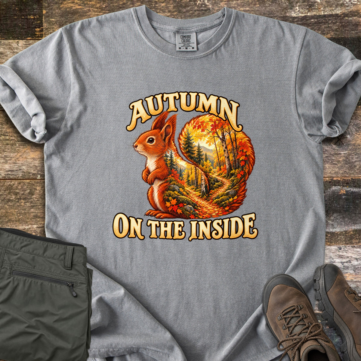 Autumn on the Inside Comfort Colors T-shirt in Grey, laid flat. A red squirrel whose tail frames a forest path through orange and yellow autumn trees, with the words Autumn on the Inside. Olive cargo pants and brown hiking boots lie beside it on weathered wood.