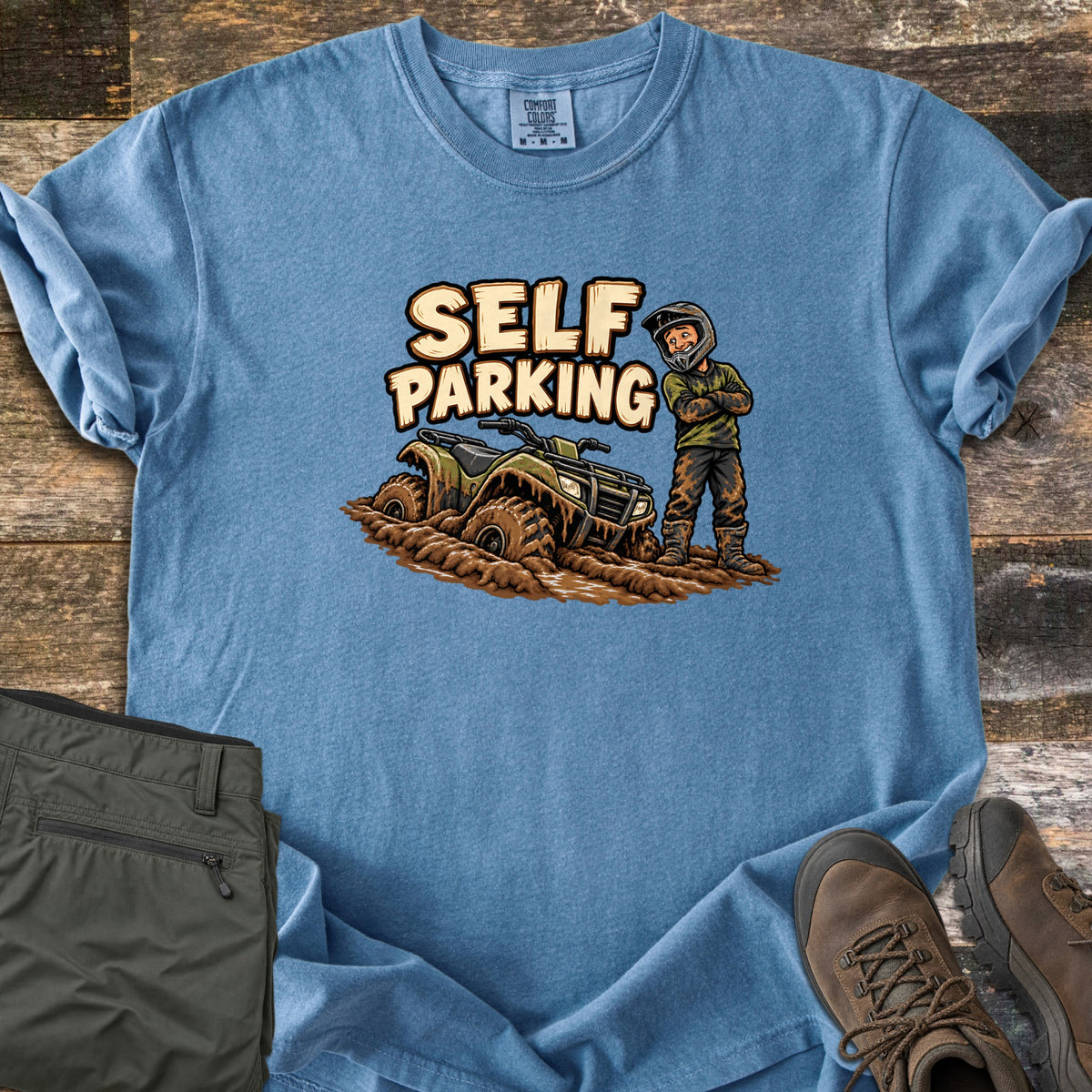 Self Parking Comfort Colors T-shirt in Blue Jean, laid flat. A cartoon rider in a helmet stands arms crossed, covered in mud, beside an ATV sunk in mud under the words Self Parking. Folded olive pants and brown hiking boots sit on a wood plank background.
