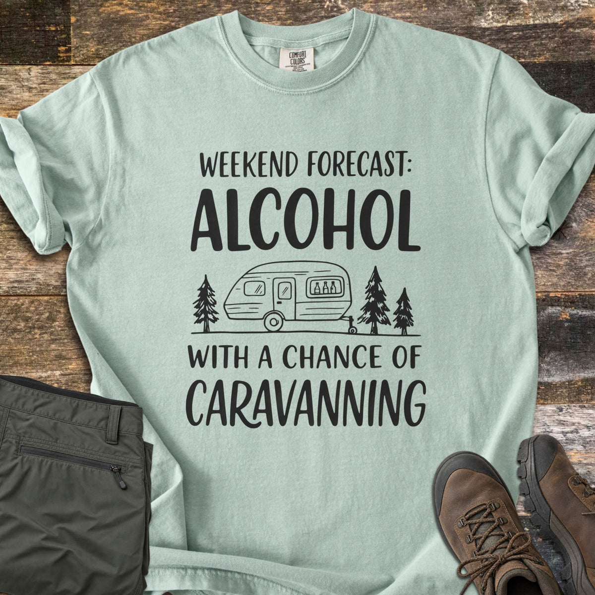 Alcohol With a Chance of Caravanning Comfort Colors T-shirt in Bay, laid flat. Black print reads Weekend Forecast: Alcohol above a caravan with bottles in its window between pine trees, then With a Chance of Caravanning. Olive trousers and brown hiking boots sit on dark wood planks.