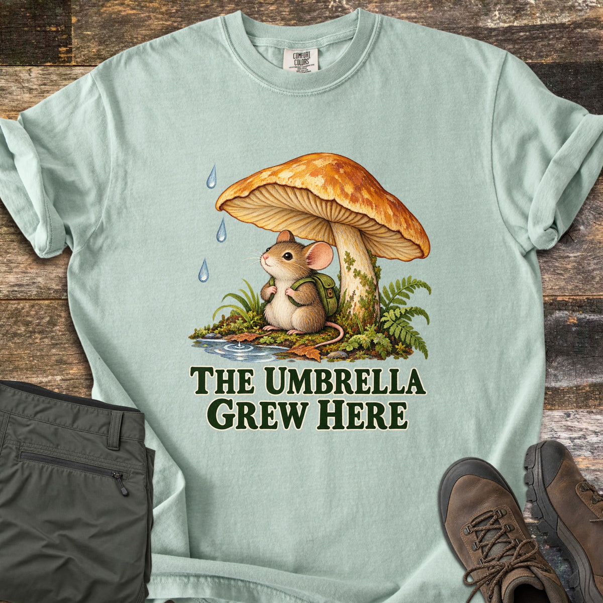 The Umbrella Grew Here Comfort Colors T-shirt in Bay, laid flat. A mouse wearing a green backpack shelters under a large orange mushroom from falling raindrops, above the words The Umbrella Grew Here. Olive pants and brown hiking boots lie beside it on weathered wood.