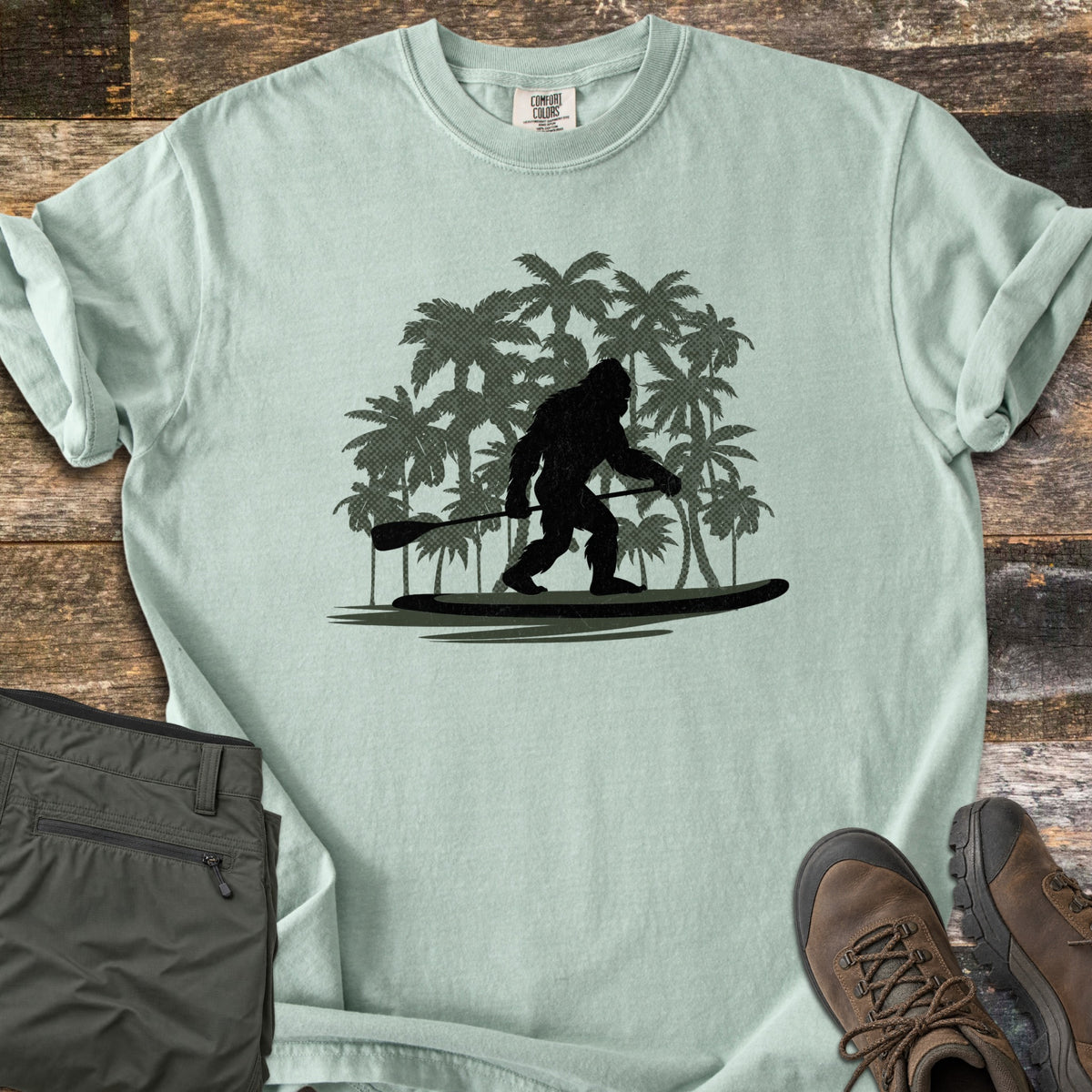 Paddle Board Squatch Comfort Colors T-shirt in Bay, laid flat. A black silhouette of Bigfoot standing on a paddle board and holding a paddle, in front of dark green palm trees. Olive pants and brown hiking boots lie beside the shirt on weathered wood planks.