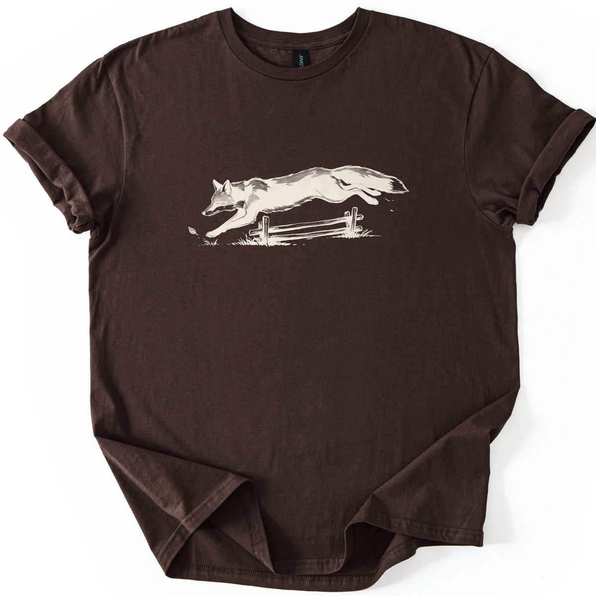 Leaping Fox Comfort Colors T-shirt in Dark Chocolate, laid flat. A cream-colored sketch of a fox leaping over a low wooden rail fence, with grass tufts and a falling leaf below. The shirt has cuffed sleeves and lies on a white background.