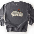 Fresh Corduroy Comfort Colors crewneck sweatshirt in Pepper, laid flat. A rider in a red jacket and helmet drives a snowmobile over groomed corduroy snow lines past two pine trees, above the words Fresh Corduroy in cream lettering. The sweatshirt lies on a white surface.