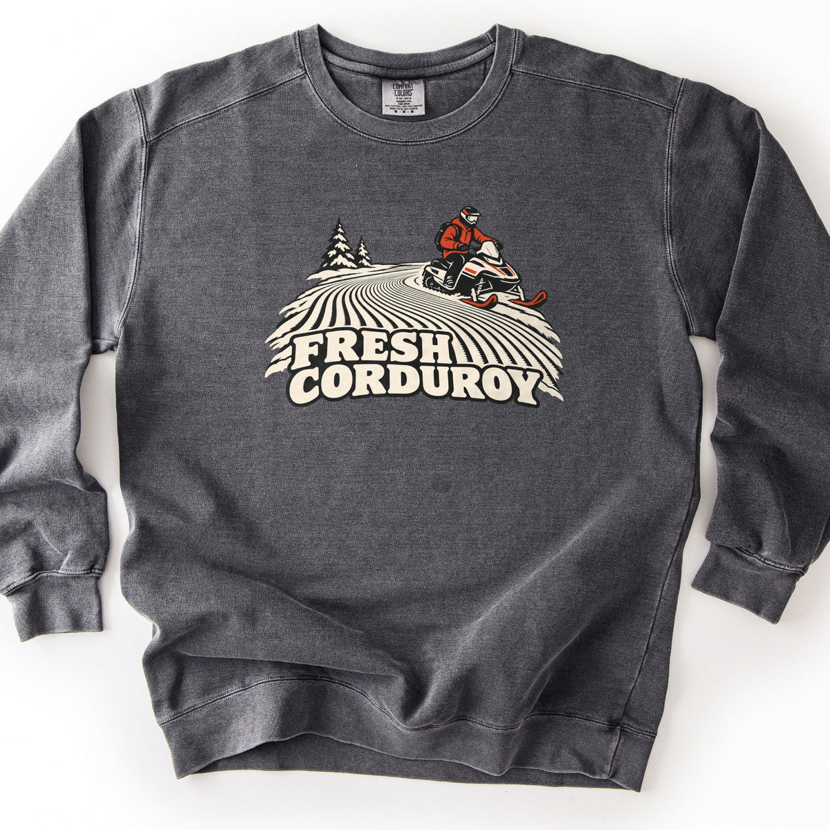 Fresh Corduroy Comfort Colors crewneck sweatshirt in Pepper, laid flat. A rider in a red jacket and helmet drives a snowmobile over groomed corduroy snow lines past two pine trees, above the words Fresh Corduroy in cream lettering. The sweatshirt lies on a white surface.
