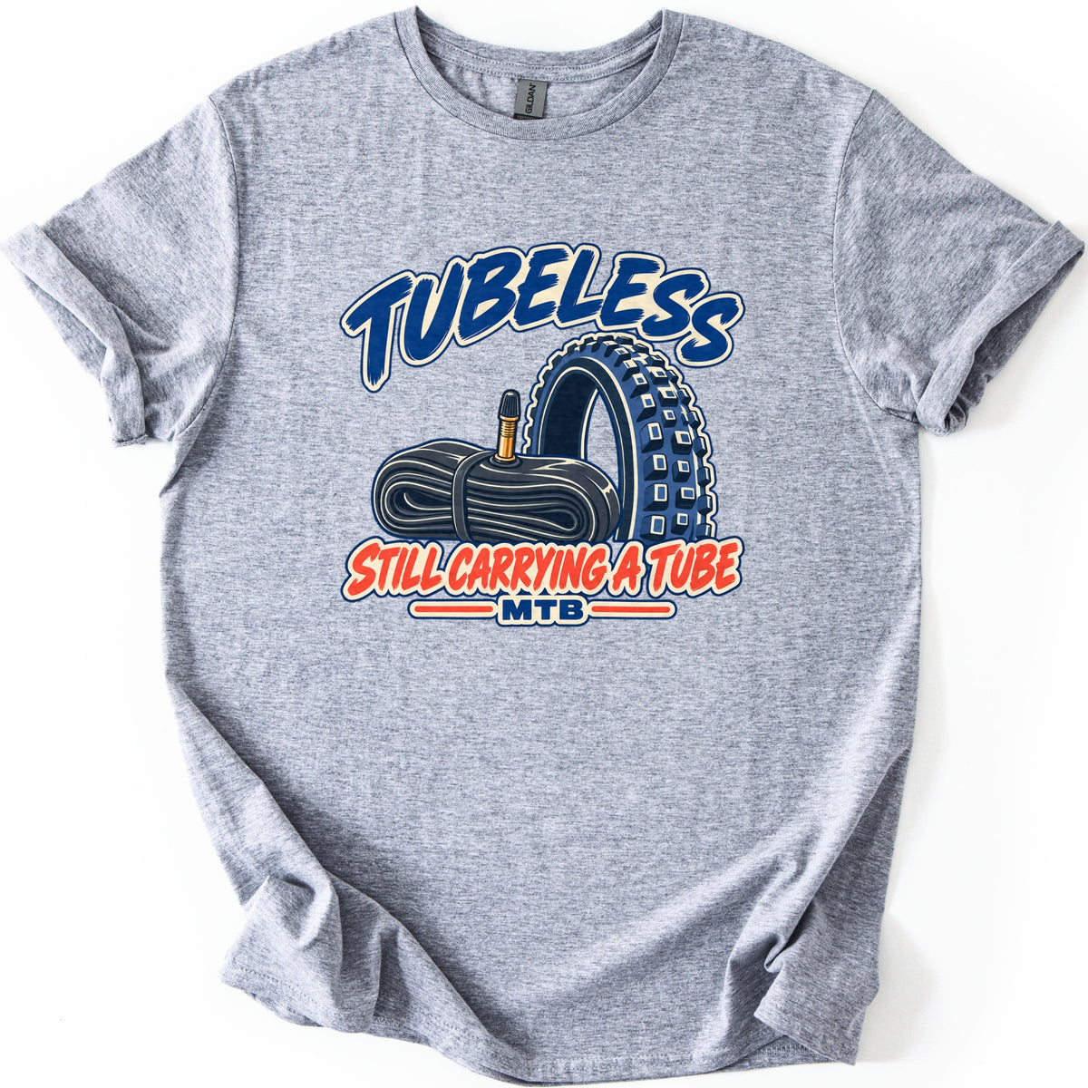 Tubeless Still Carrying a Tube Comfort Colors T-shirt in Sport Grey, laid flat. A rolled-up black inner tube with a brass valve sits beside a knobby blue mountain bike tire, between the words Tubeless and Still Carrying a Tube, with MTB below. Sleeves are cuffed on a white background.
