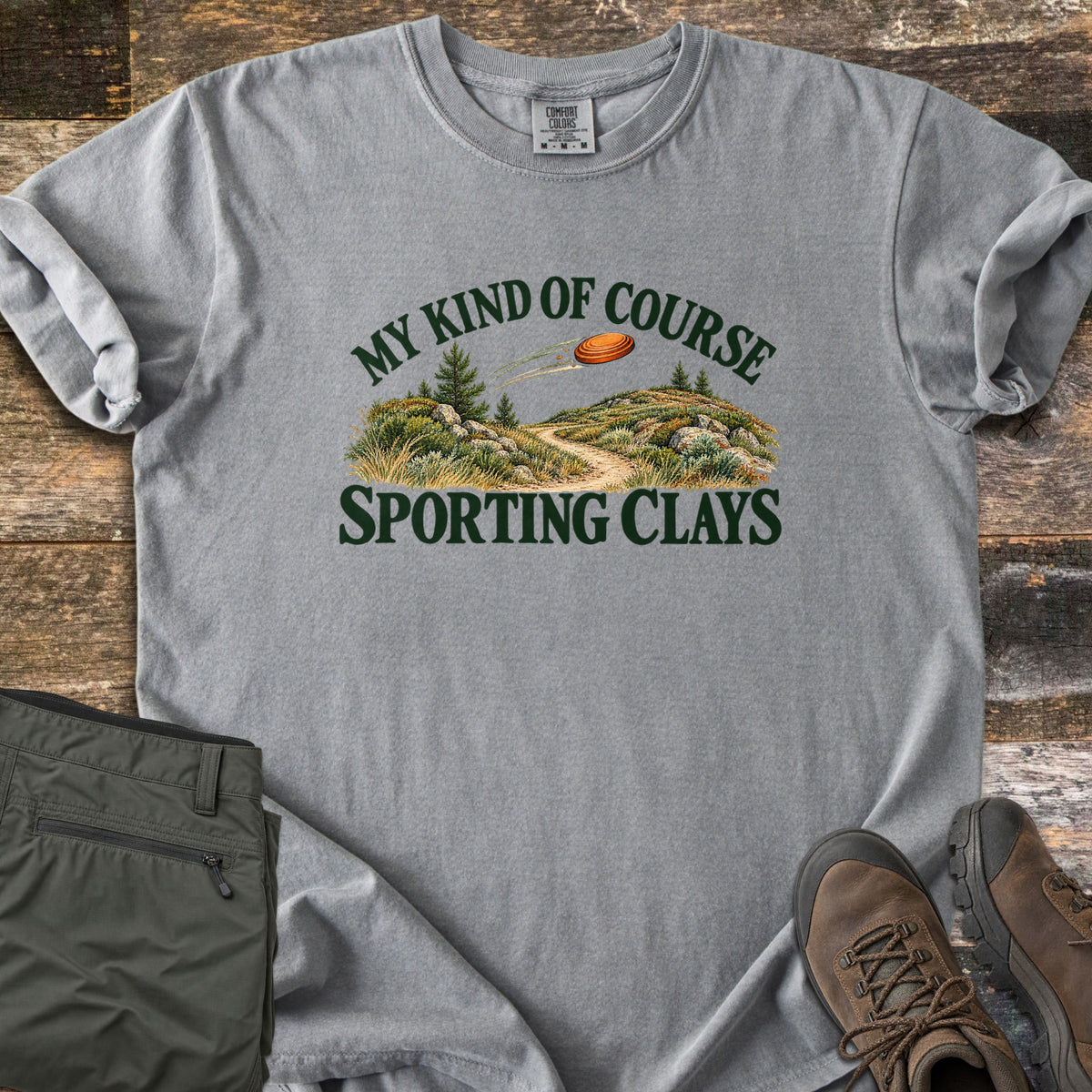 My Kind of Course Comfort Colors T-shirt in Grey, laid flat. The words My Kind of Course arch over a painted trail winding between grassy, rocky hills with pine trees as an orange clay target flies past, above Sporting Clays. Olive pants and brown hiking boots sit on weathered wood.