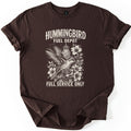 Hummingbird Fuel Depot Comfort Colors T-shirt in Dark Chocolate, laid flat. A cream etched-style hummingbird sips from a hanging nectar feeder among large flowers, between the words Hummingbird Fuel Depot and Full Service Only. The sleeves are rolled on a plain white background.