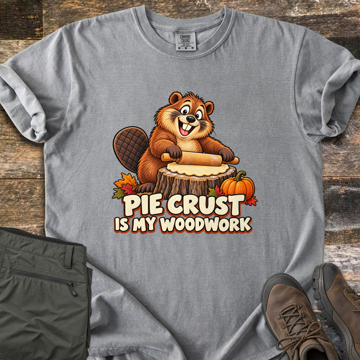 Pie Crust Is My Woodwork Comfort Colors T-shirt in Grey, laid flat. A grinning cartoon beaver rolls pie dough on a tree stump, with a small pumpkin and autumn leaves, above the words Pie Crust Is My Woodwork. Olive pants and brown hiking boots sit on a rustic wood surface.