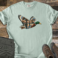 Mallard Flooded Oaks Comfort Colors T-shirt in Bay, laid flat. A flying mallard drake whose wings and body are filled with a scene of moss-draped oaks standing in still water at sunset. Laid on weathered wood beside folded olive pants and brown hiking boots.