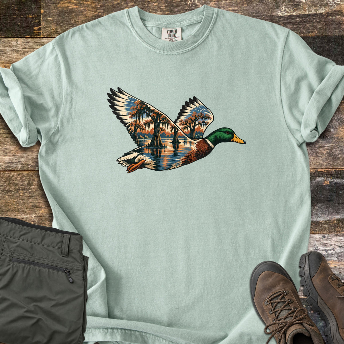 Mallard Flooded Oaks Comfort Colors T-shirt in Bay, laid flat. A flying mallard drake whose wings and body are filled with a scene of moss-draped oaks standing in still water at sunset. Laid on weathered wood beside folded olive pants and brown hiking boots.