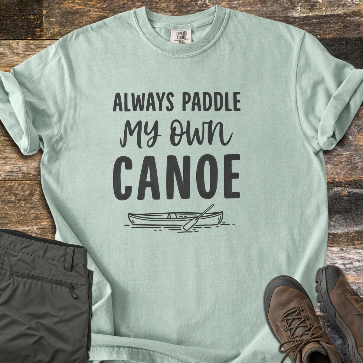 Paddle My Own Canoe Comfort Colors T-shirt in Bay, laid flat. Charcoal text reads Always Paddle My Own Canoe above a line drawing of a canoe with a paddle on the water. It lies on weathered wood with rolled sleeves, folded olive hiking pants, and brown hiking boots.