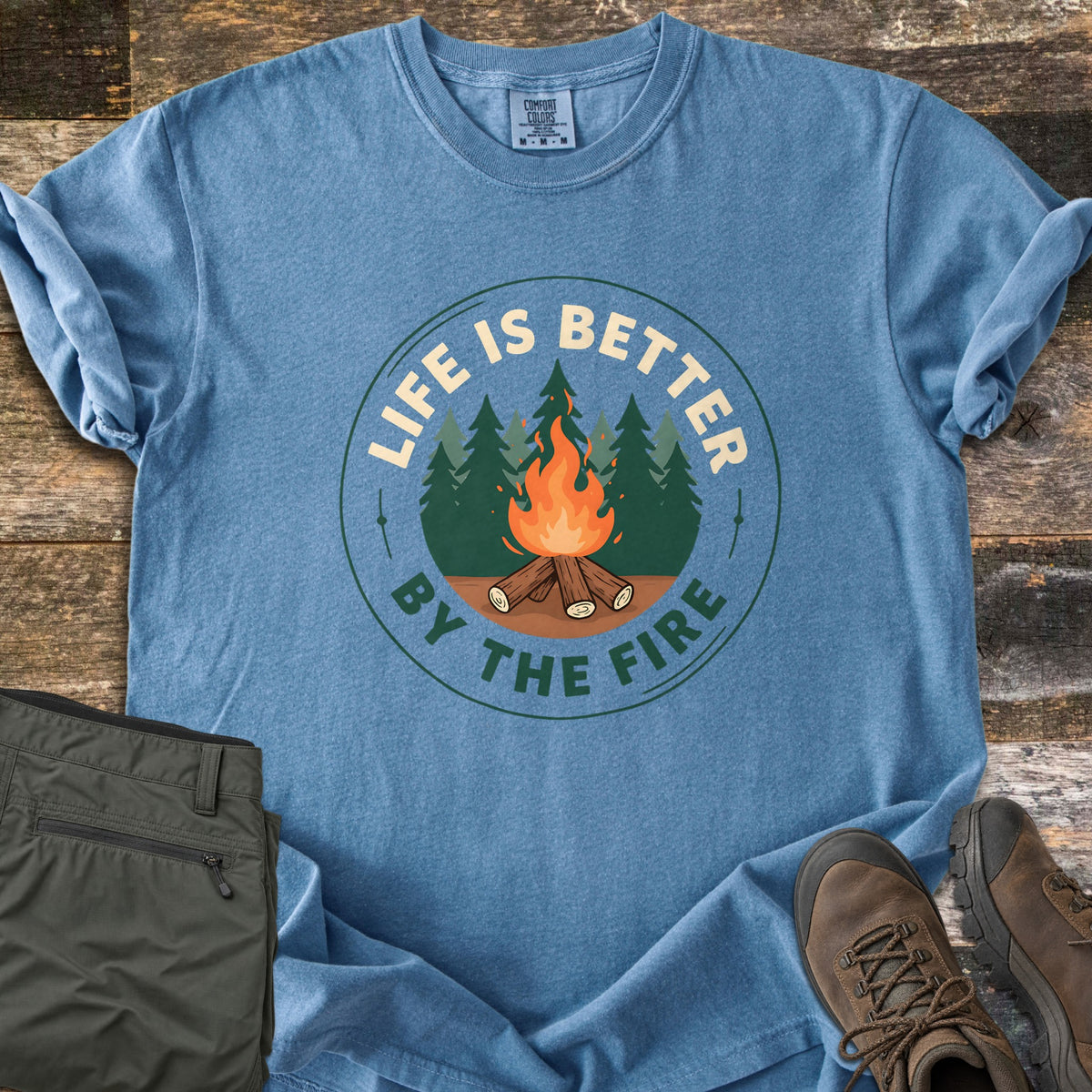 Life Is Better By The Fire Badge Comfort Colors T-shirt in Blue Jean, laid flat. A round badge shows a campfire of crossed logs before pine trees, with LIFE IS BETTER arched above and BY THE FIRE below. Olive hiking pants and brown hiking boots sit beside it on weathered wood planks.