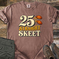 25 Straight Skeet Comfort Colors T-shirt in Espresso, laid flat. Large cream numbers 25 with two orange clay targets flying beside them, above the words Straight Skeet in gold and cream. Sleeves are rolled on a wood plank surface with olive cargo pants and brown hiking boots.