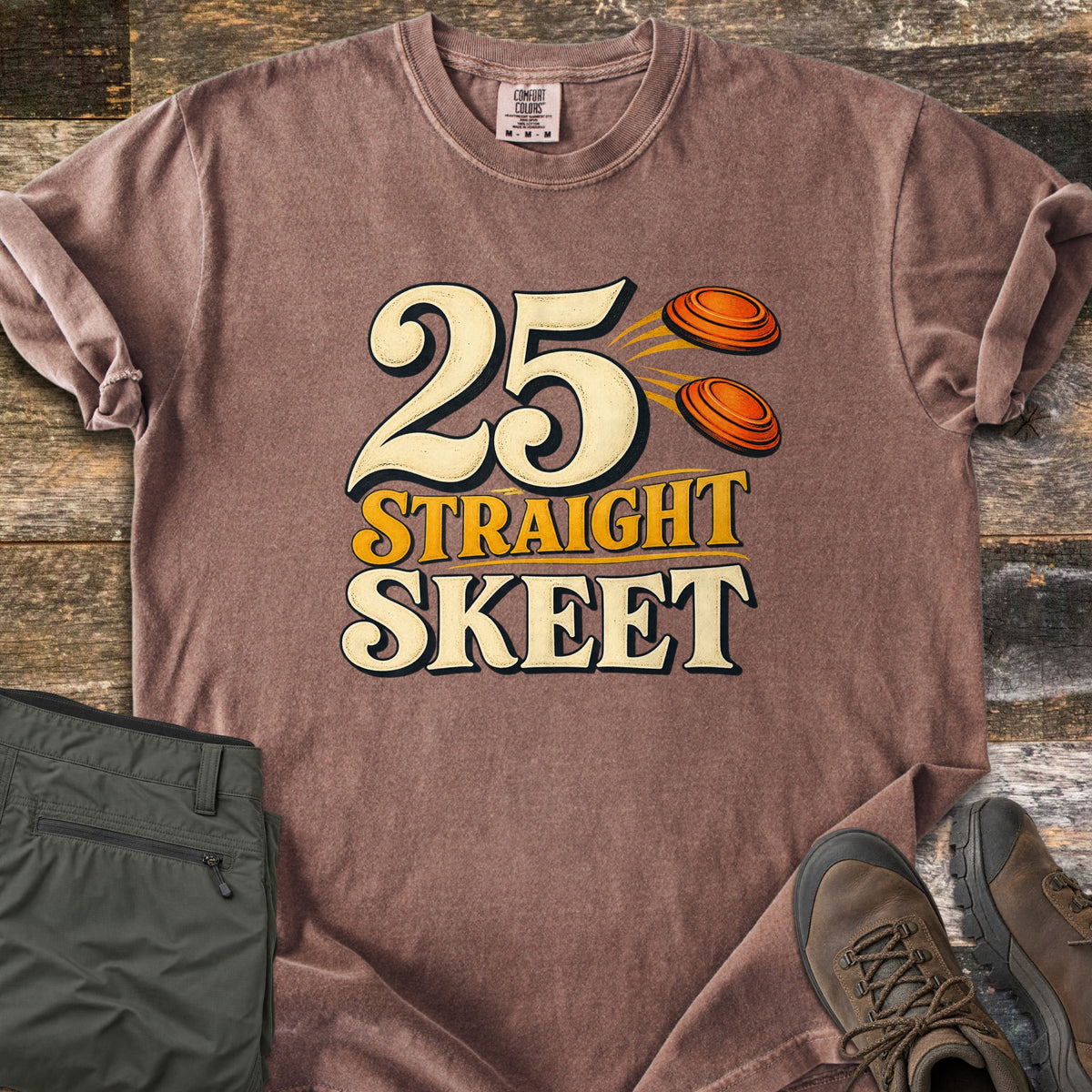 25 Straight Skeet Comfort Colors T-shirt in Espresso, laid flat. Large cream numbers 25 with two orange clay targets flying beside them, above the words Straight Skeet in gold and cream. Sleeves are rolled on a wood plank surface with olive cargo pants and brown hiking boots.