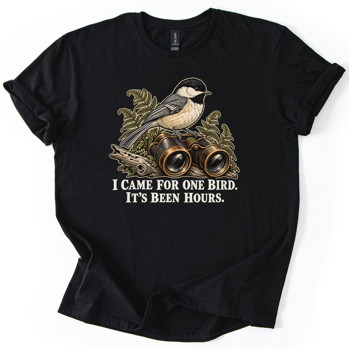 I Came for One Bird. It's Been Hours. Comfort Colors T-shirt in Black, laid flat. A black-capped chickadee perches on brass binoculars resting on a weathered log among green ferns, above the words I Came for One Bird. It's Been Hours. The sleeves are rolled on a white background.
