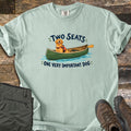 Important Dog Comfort Colors T-shirt in Bay, laid flat. A smiling golden retriever in an orange life vest sits in a green canoe with a paddle, between the words Two Seats and One Very Important Dog. Olive hiking pants and brown boots lie on weathered wood.