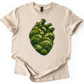 Moss Heart Comfort Colors T-shirt in Sand, laid flat. An anatomical heart made of clumps of green moss, with pale tufts, small sprouting stalks and three tube-like vessels on top, with no words. The shirt lies on a white surface with sleeves cuffed.