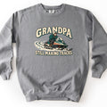 Grandpa Still Making Tracks Comfort Colors crewneck sweatshirt in Grey, laid flat. Front print shows a white-bearded grandpa in a helmet, sunglasses and green jacket riding a vintage snowmobile through curving snow tracks, between the words Grandpa and Still Making Tracks. Shown on a white background.