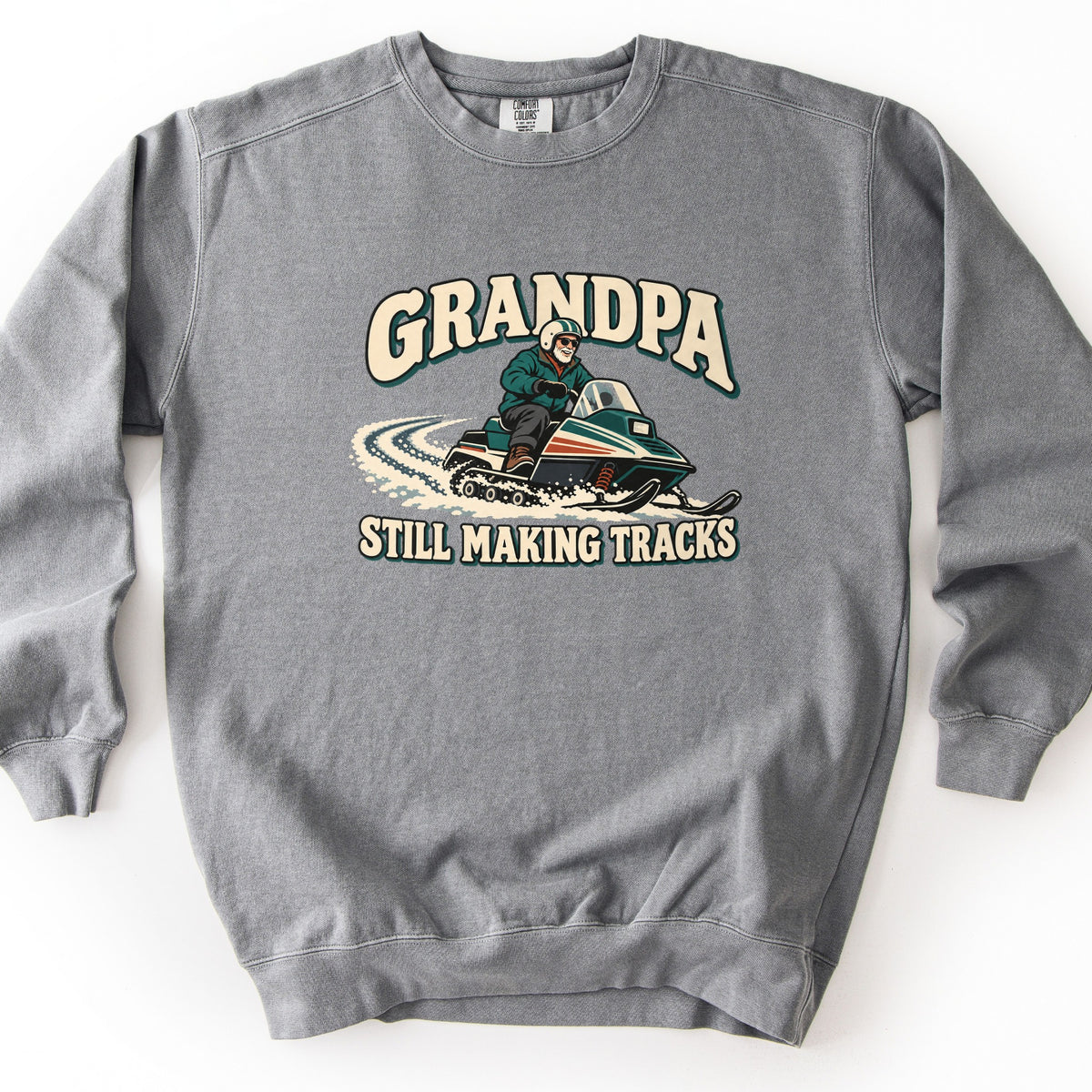Grandpa Still Making Tracks Comfort Colors crewneck sweatshirt in Grey, laid flat. Front print shows a white-bearded grandpa in a helmet, sunglasses and green jacket riding a vintage snowmobile through curving snow tracks, between the words Grandpa and Still Making Tracks. Shown on a white background.
